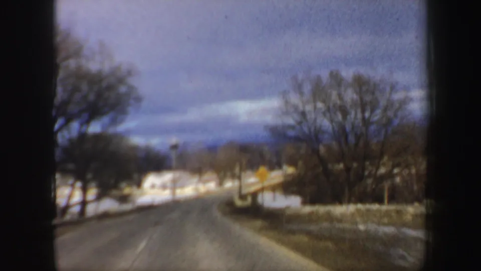 1962 New Hampshire 8mm frame looking down a winter road toward a small light-colored house among bare trees and snow patches.