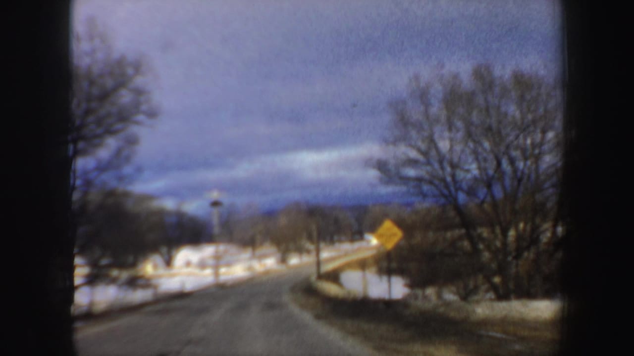 Snow-lined rural road curving past bare trees under a cloudy sky.