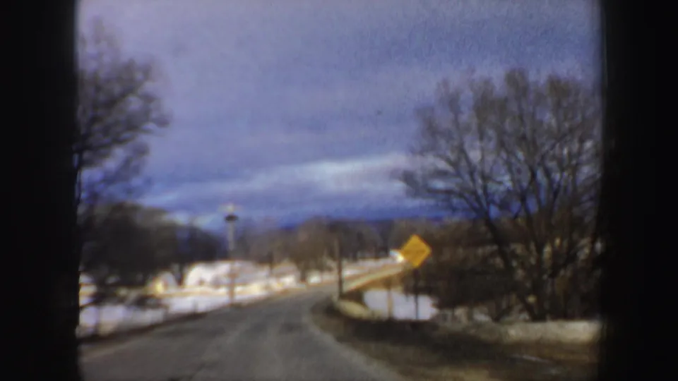 1962 New Hampshire 8mm view from a car window of a snow-lined rural road, bare trees, and a yellow warning sign.