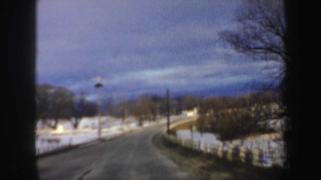 Winter road bending right with snowbanks, trees, and a distant house-like structure.