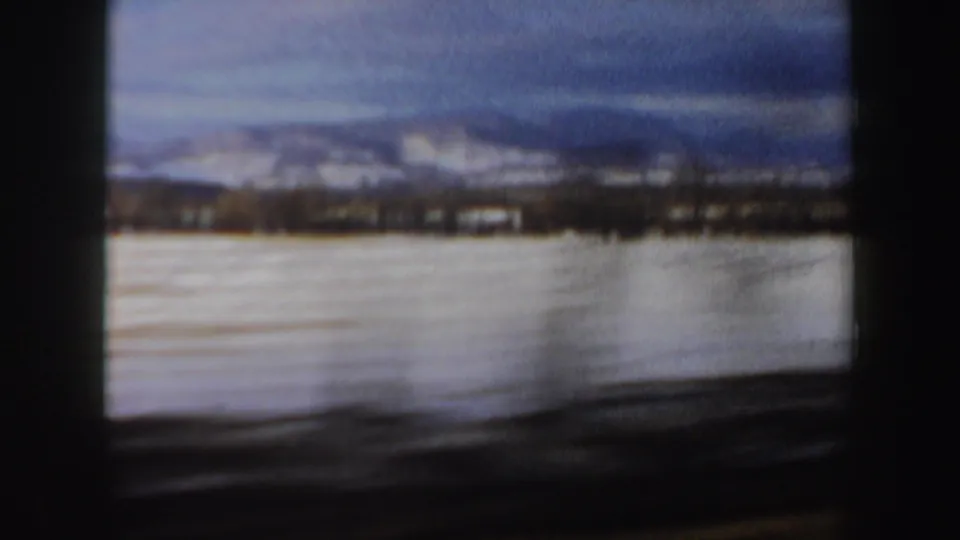 1962 New Hampshire winter landscape with a snow-covered field, distant tree line, hills, and cloudy blue sky from a moving car.
