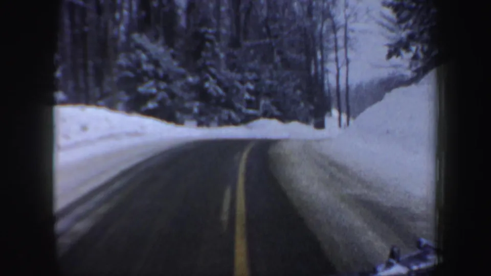 Late-frame view of the 1962 New Hampshire drive, with the road curving beside snowbanks, evergreens, bare trees, and dark 8mm frame edges.