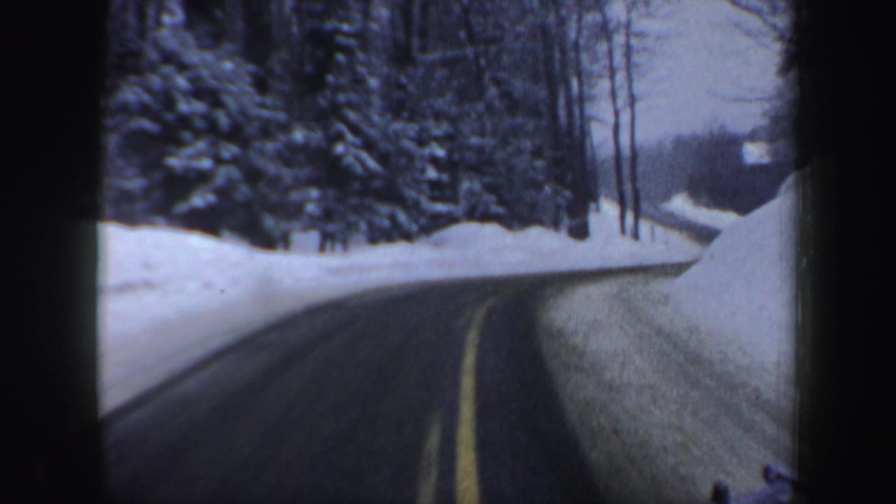 1962 New Hampshire 8mm frame showing a wet road with a yellow center line between plowed snowbanks and snowy trees.