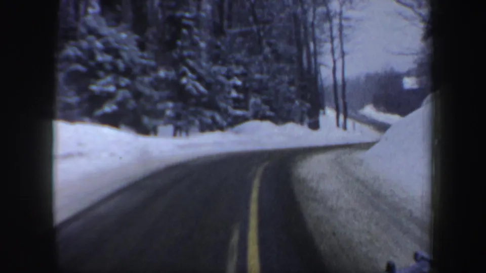 1962 New Hampshire 8mm frame showing a wet road with a yellow center line between plowed snowbanks and snowy trees.
