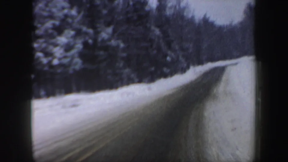 1962 New Hampshire winter driving frame with a narrow road, snow piled along the shoulder, and dense trees beyond.