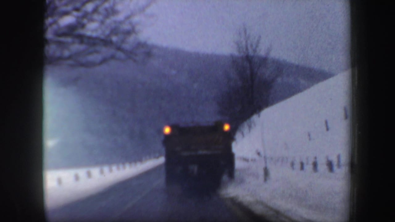 Snow plow ahead on a wet road between high snowbanks and wooded hills.