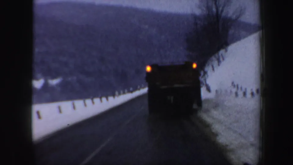 1962 New Hampshire home-movie view of a snow plow ahead on a curving road, with snowbanks and winter hills in the background.