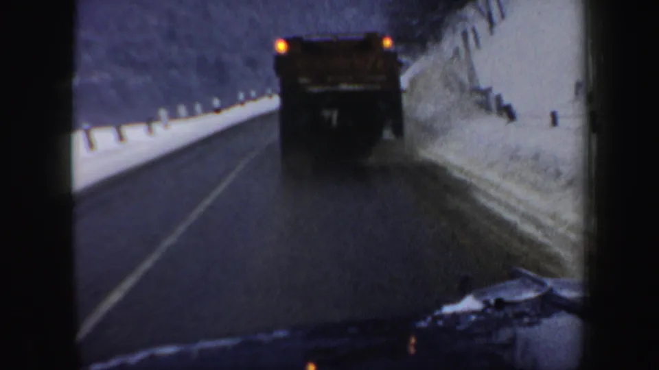 1962 New Hampshire 8mm frame of a plow driving ahead on a slick-looking road, with snowbanks, roadside posts, and the filming vehicle edge visible.