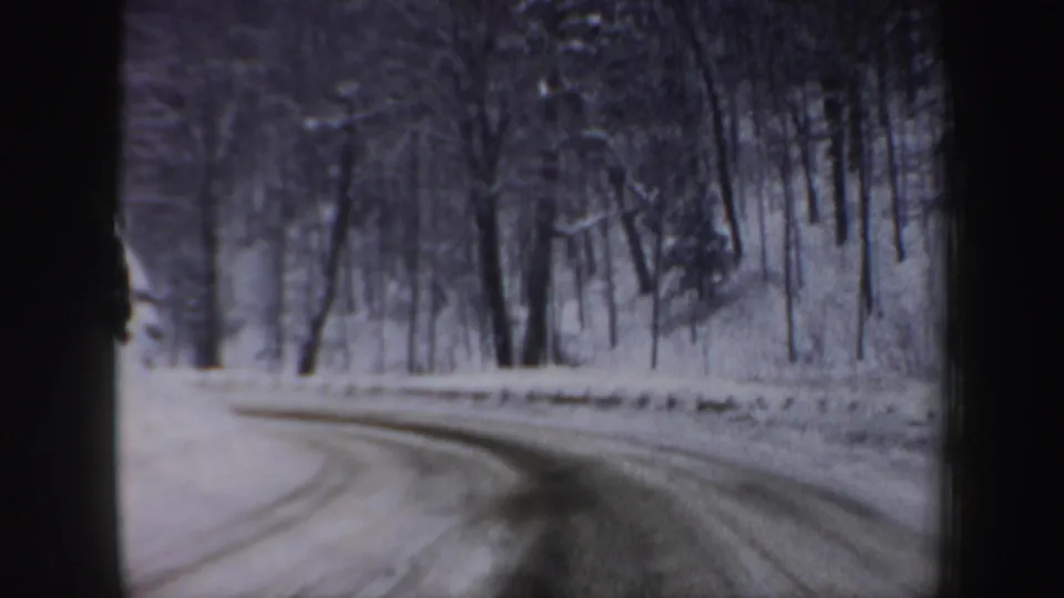 Vehicle-view 8mm frame of a snow-covered New Hampshire forest road bending right in winter daylight.