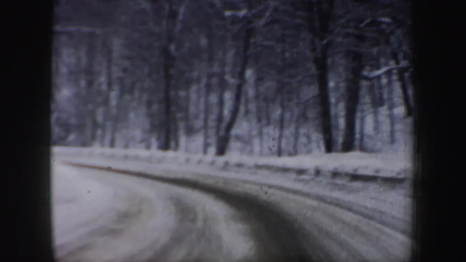 1960s New Hampshire winter driving view with a packed snowy road, bare trees, and soft 8mm grain.