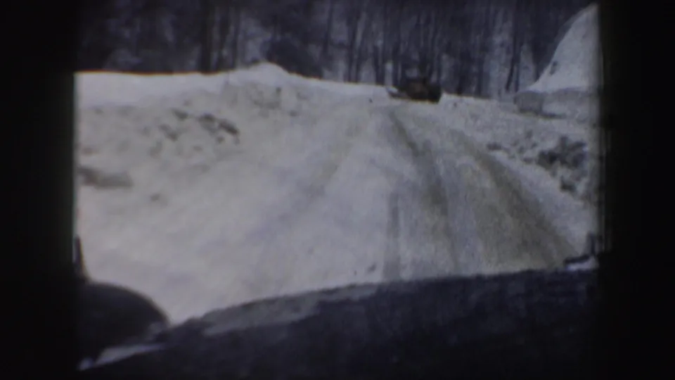 New Hampshire winter road scene from 1962 home-movie footage, showing snowbanks and a dark vehicle ahead.