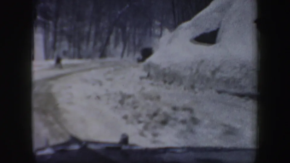 In-car 8mm frame from 1962 New Hampshire showing a snowbank beside a wooded road, with a small distant figure or object ahead.