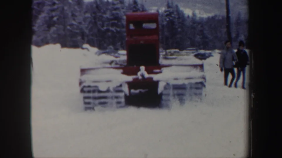 1962 New Hampshire 8mm frame showing a red tracked snow vehicle from the front as two distant onlookers stand in the snow.