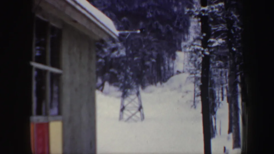1962 New Hampshire home-movie frame showing a resort building edge, lift tower, snowy ground, and forest backdrop.