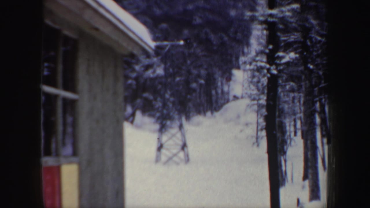 1962 New Hampshire home-movie frame showing a snowy ski-area view through a building edge toward trees and a lift tower.