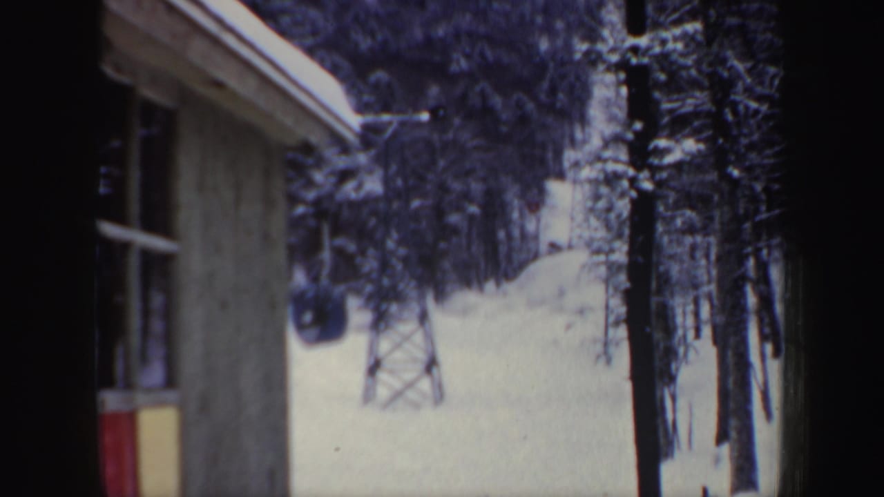 Snow-covered hillside with a lift tower and trees seen beside a building wall.