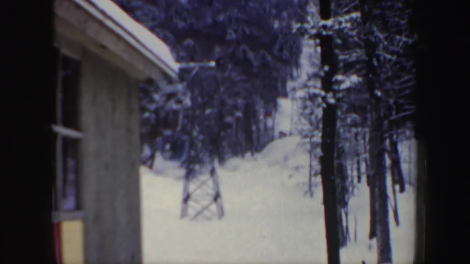 A 1962 home-movie frame looks past a wooden building toward a snowy New Hampshire ski lift area and forest.