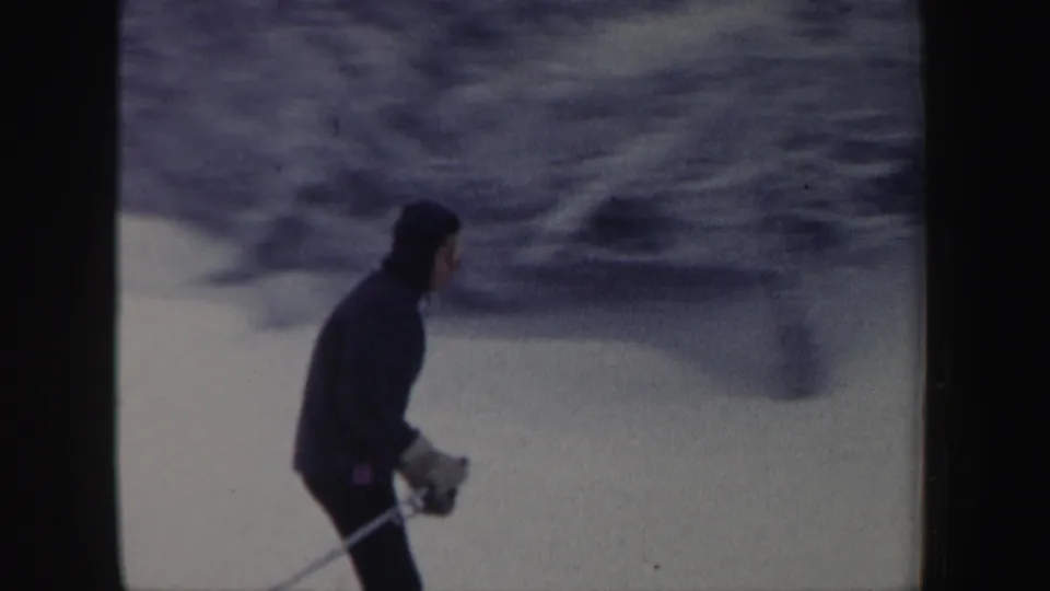 A skier in a dark hooded jacket and ski poles crosses the snowy slope in 1962 New Hampshire footage.
