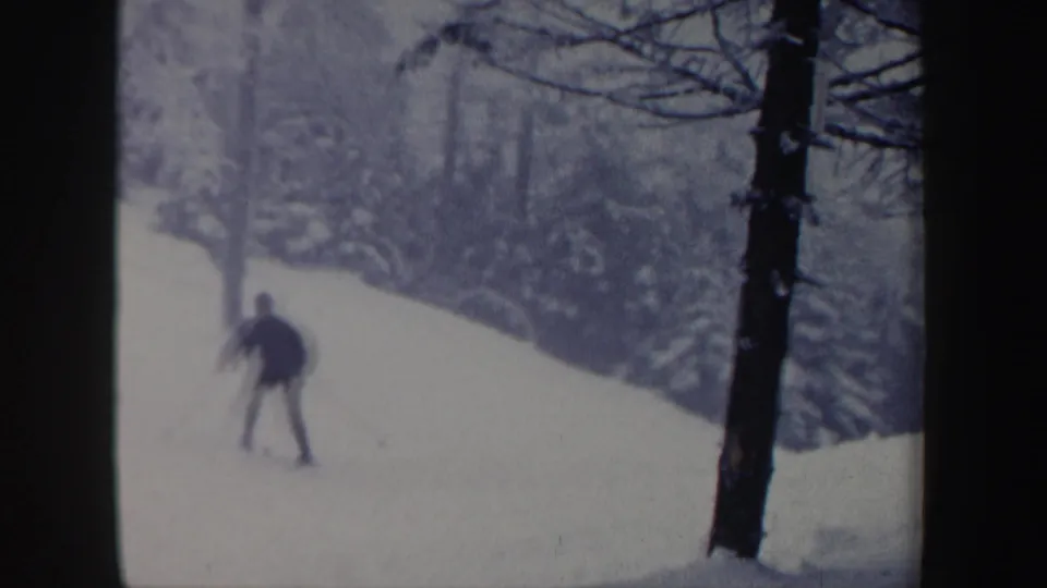 1962 New Hampshire home-movie frame of a dark-clothed skier moving downhill on a gentle snowy slope lined with winter trees.