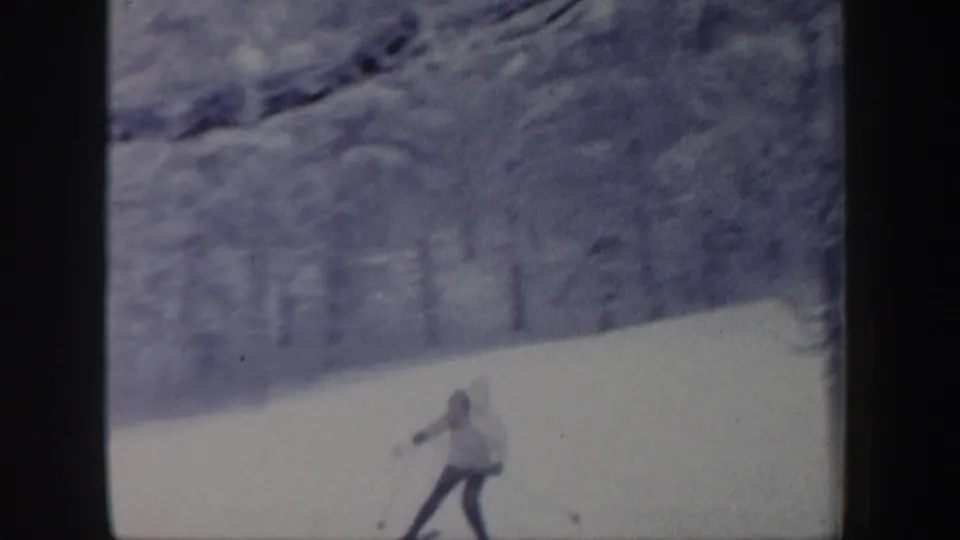 1962 New Hampshire 8mm frame with a skier in a light jacket moving across a snow-covered hillside below blurred winter trees.