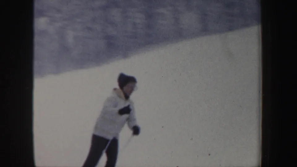 Close 1962 New Hampshire home-movie frame of a skier in a light jacket and dark knit hat using poles on a snowy slope.