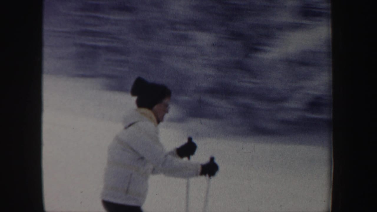 In 1962 New Hampshire home-movie footage, a skier in a white jacket and dark knit hat uses poles while moving across packed snow.