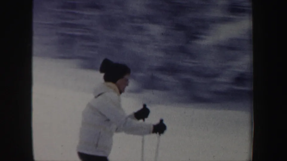 In 1962 New Hampshire home-movie footage, a skier in a white jacket and dark knit hat uses poles while moving across packed snow.