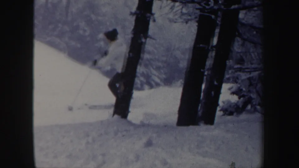 The skier moves near dark tree trunks on a snowy slope, with soft 8mm grain and winter woods visible around the figure.