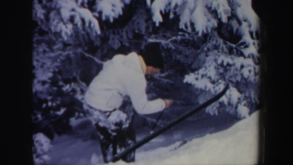 The skier slows or stops near snow-laden evergreen branches, bending forward with skis and poles visible in the winter scene.