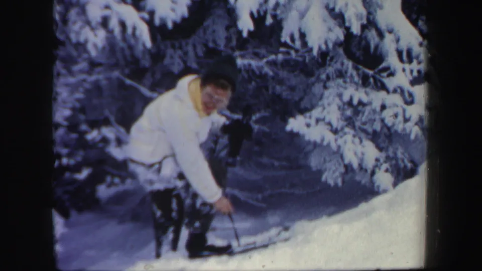 A woman in a white jacket and dark pants leans into the snowy climb, framed by heavy evergreen branches.