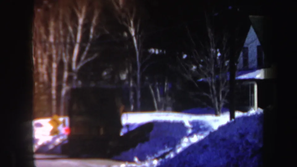 Dark 8mm frame of a snowy New Hampshire residential street with a truck-like vehicle, hydrant, bare trees, and house exterior.