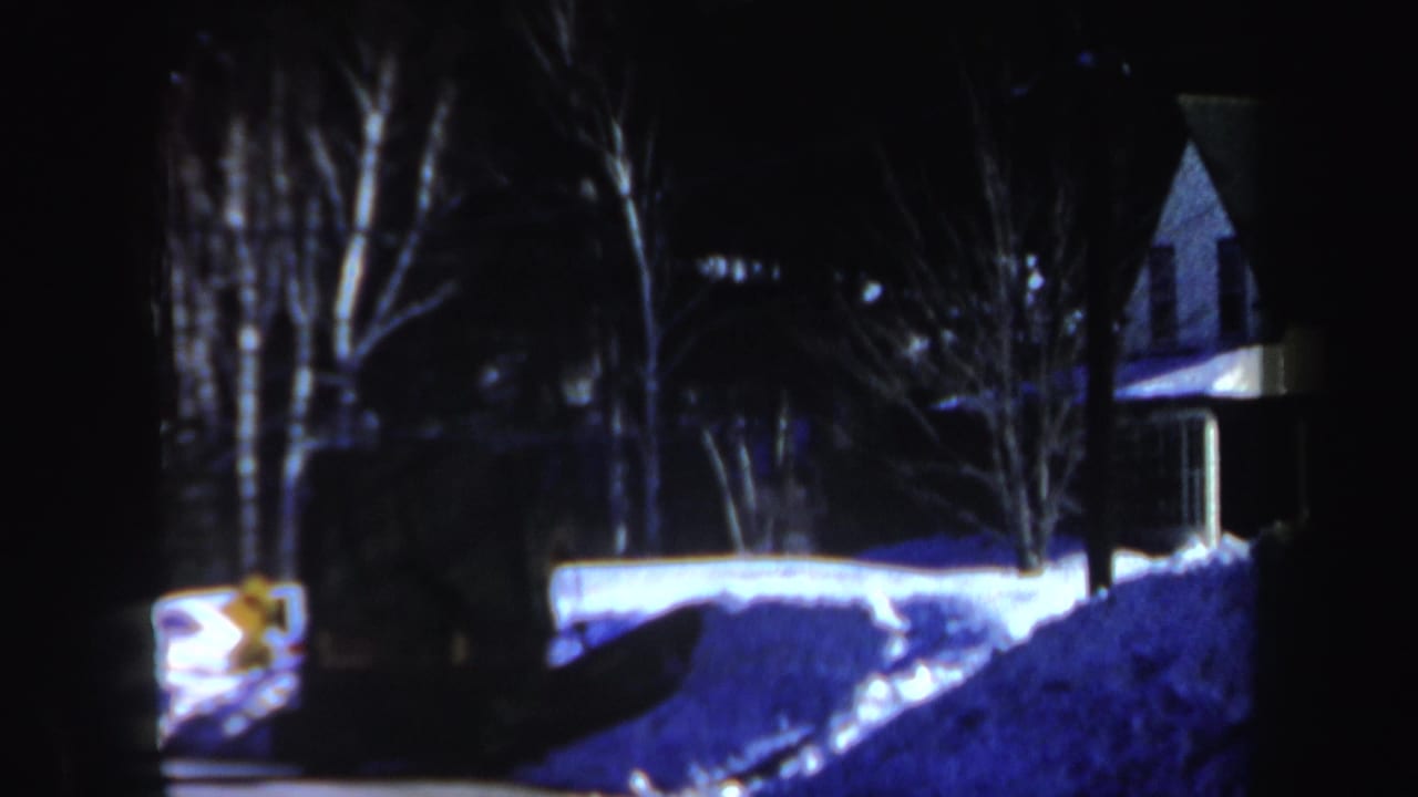 1962 New Hampshire 8mm frame showing a dark snowy roadside, bare trees, a house exterior, and a glowing car light near a snowbank.