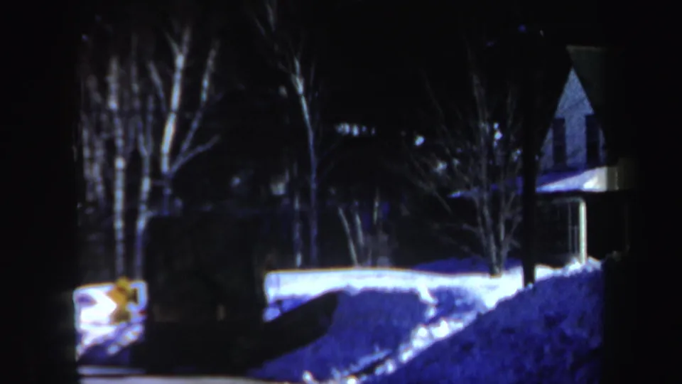 1962 New Hampshire 8mm frame showing a dark snowy roadside, bare trees, a house exterior, and a glowing car light near a snowbank.