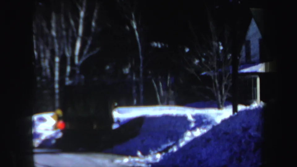 1962 New Hampshire winter frame with a car's red light beside high snowbanks, bare trees, and a small house exterior in low light.