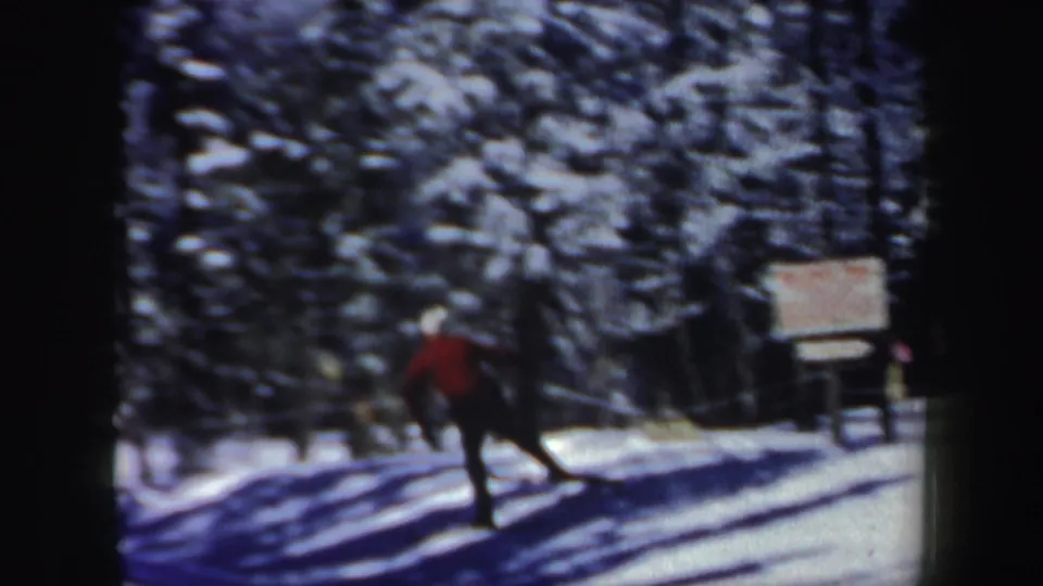 In 1962 New Hampshire footage, a skier in a red jacket travels down a snowy forest path with a trail sign ahead.