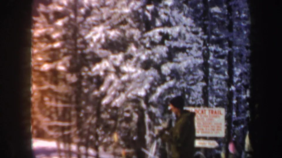 A person in dark winter clothing stands beside a Wildcat Trail sign among snow-covered evergreens in 1962 New Hampshire footage.