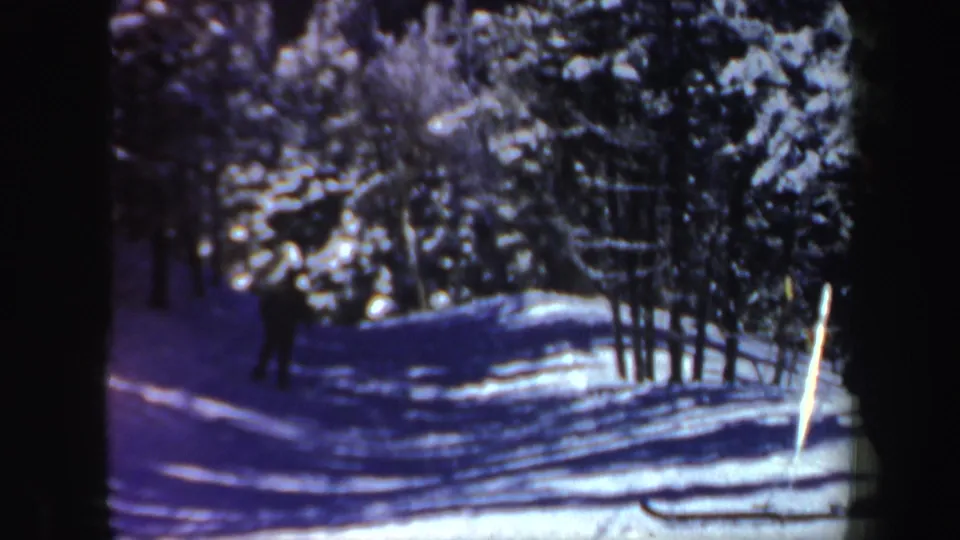 A distant skier travels through a snowy evergreen corridor, with cool blue shadows and soft 8mm color.