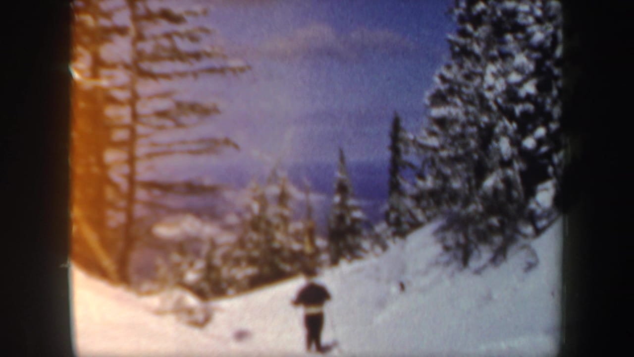 A small figure skis across a snowy New Hampshire mountain slope framed by tall pines and distant winter hills.