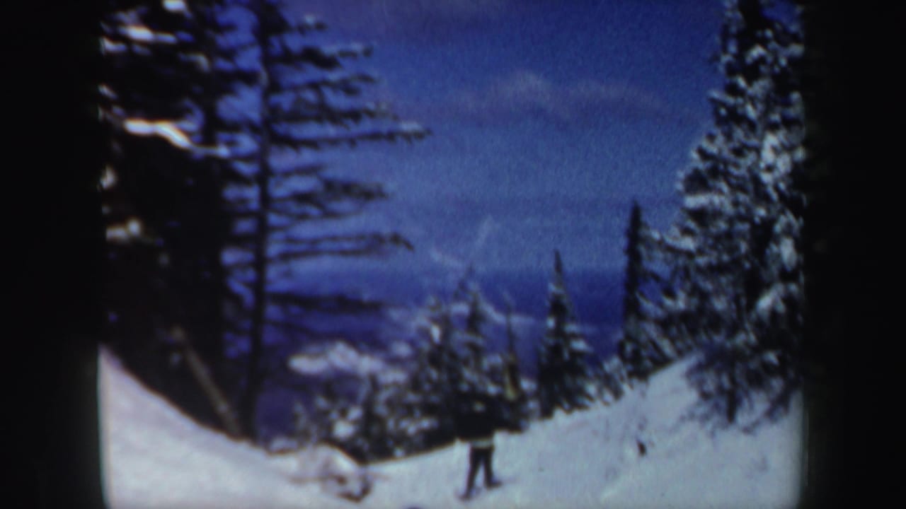1962 New Hampshire home-movie frame showing a distant skier crossing a snow-covered forest slope under a blue winter sky.