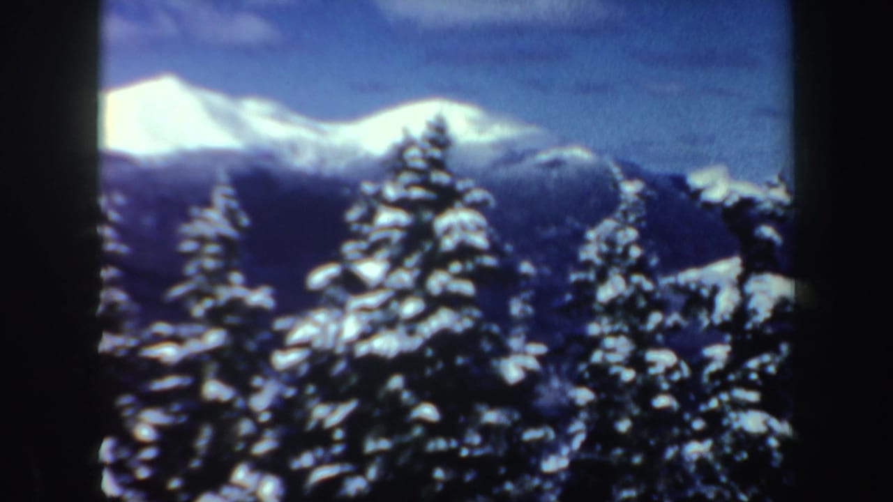 Wide 8mm frame of New Hampshire snow-covered pines and distant bright mountain ridges, with any skier indistinct or out of view.