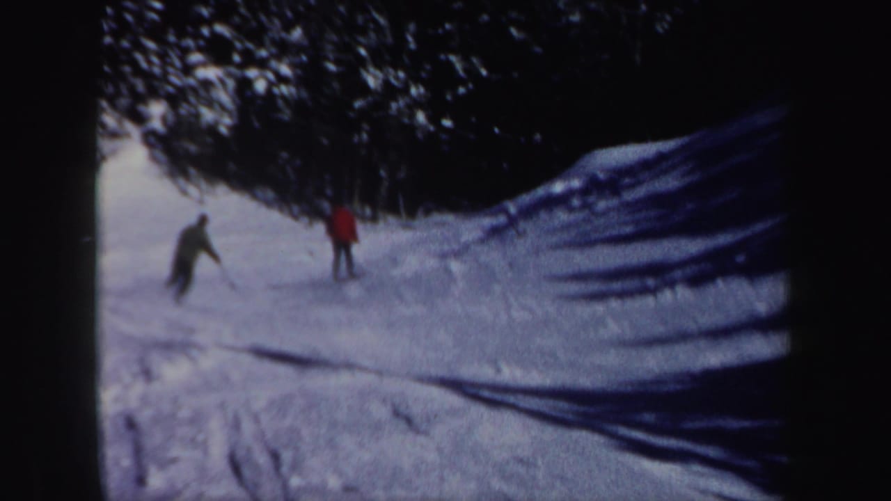 Two men practice on a snowy New Hampshire slope in 1962, with one wearing a red coat against the dark tree line.