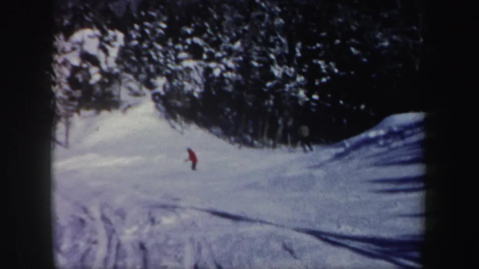 1962 New Hampshire 8mm frame showing a distant skier in red crossing a sunlit snowy slope near evergreen trees.