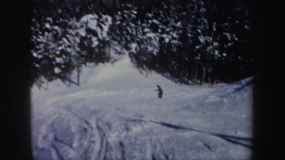 1962 New Hampshire home-movie frame with a distant skier descending a snowy run edged by evergreen trees.