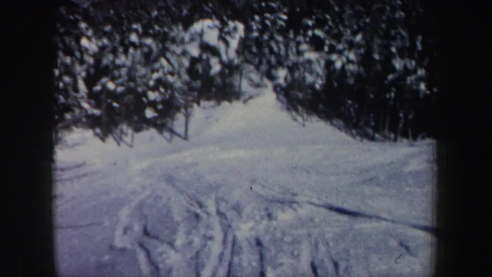 1962 New Hampshire 8mm frame of a snowy ski slope marked by tracks and bordered by evergreen trees.