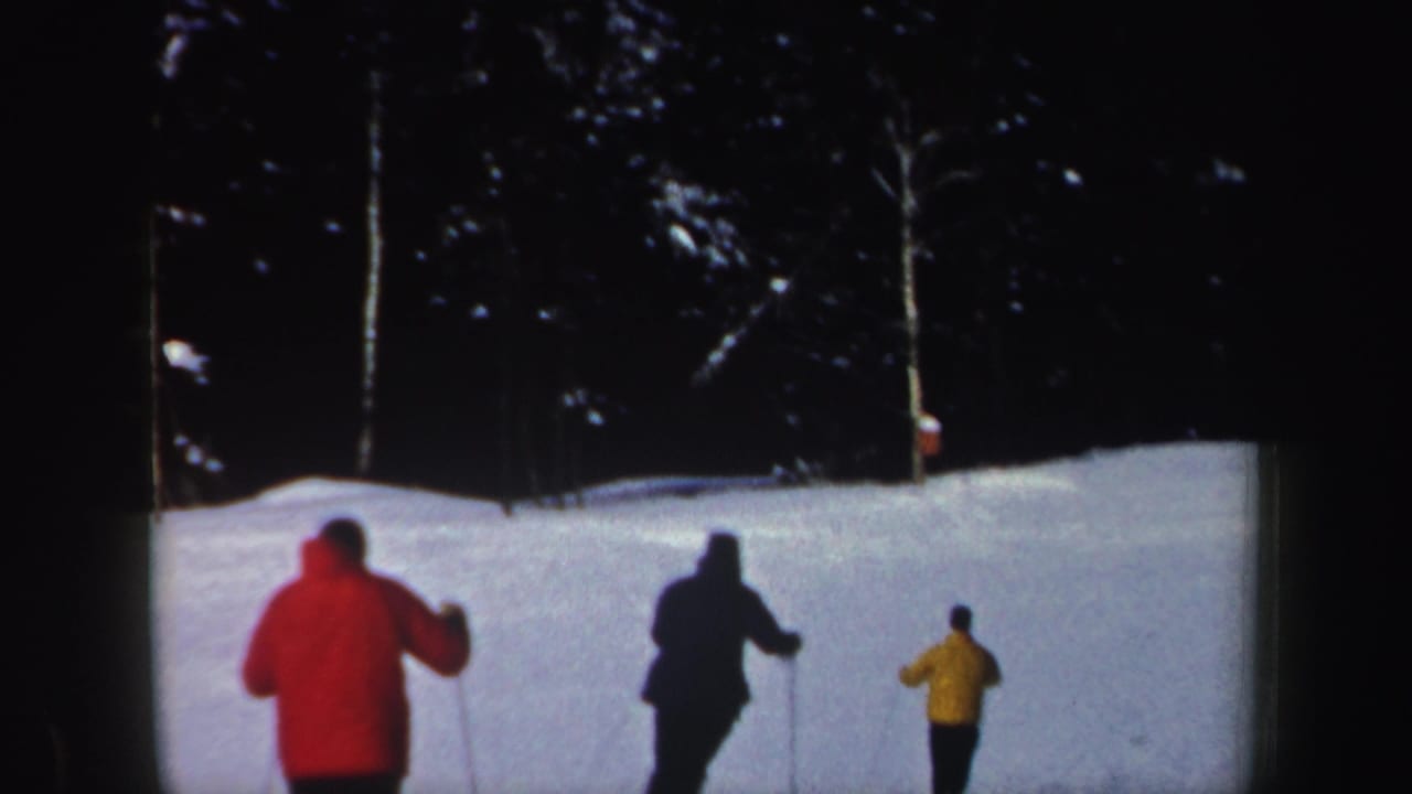Three adult skiers in red, dark, and yellow outerwear move across a snowy New Hampshire slope in the 1962 home movie.