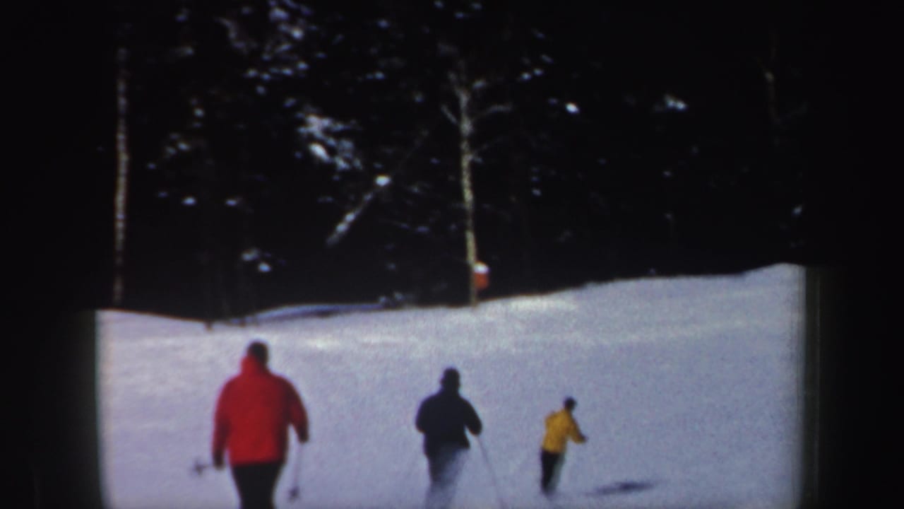 Three skiers in red, dark, and yellow outerwear descend a snowy New Hampshire slope in 1962 home-movie footage.