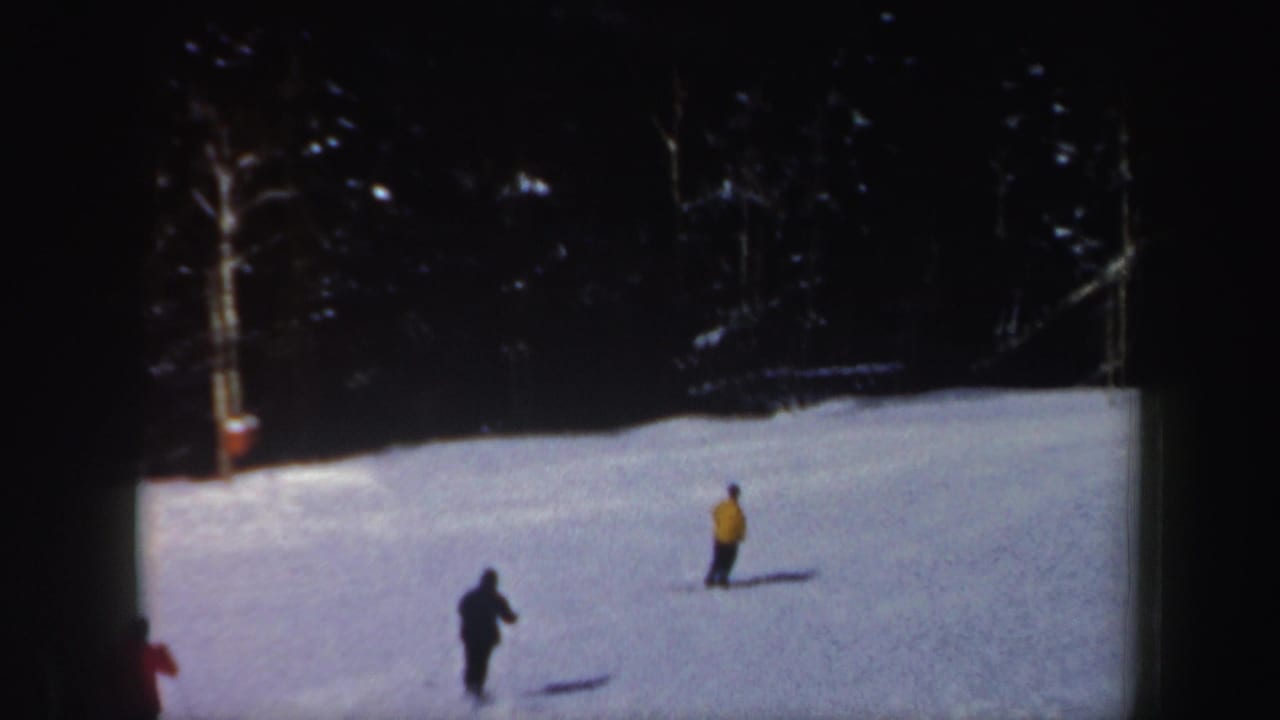 Two skiers in dark and yellow outerwear continue across the snowy New Hampshire slope as another skier exits the edge of frame.