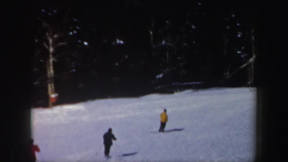 Two skiers in dark and yellow outerwear continue across the snowy New Hampshire slope as another skier exits the edge of frame.