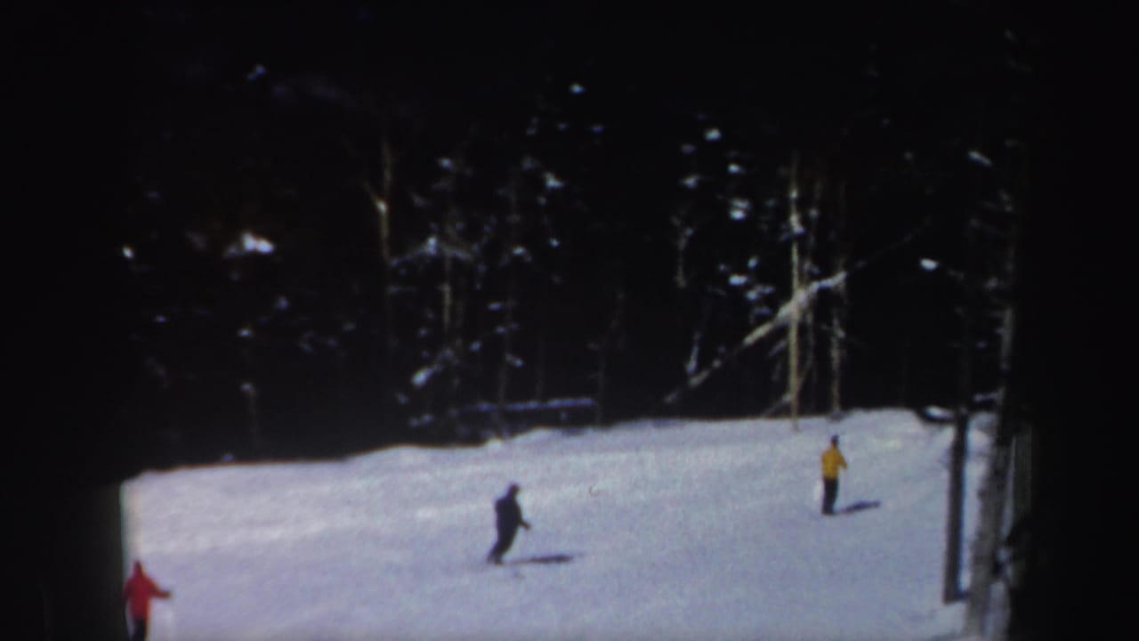Two distant skiers cross a snowy wooded slope in 1962 New Hampshire home-movie footage, with a red figure partly visible at left.