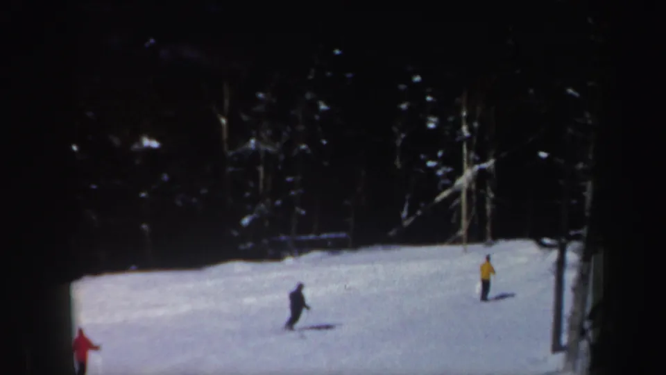 Two distant skiers cross a snowy wooded slope in 1962 New Hampshire home-movie footage, with a red figure partly visible at left.